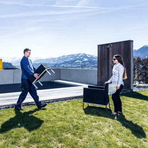 Zwei Personen tragen Büromöbel vor einer Berglandschaft